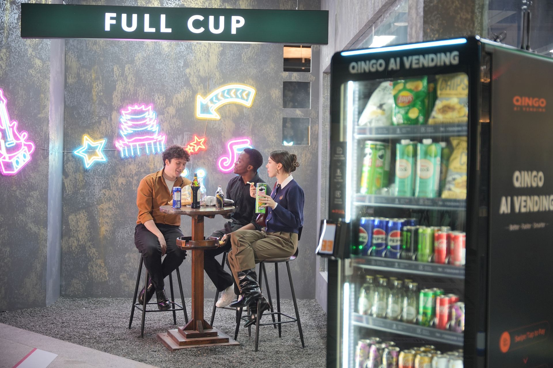 Black vending machine on a city sidewalk, stocked with snacks and drinks, orange cylinder nearby, modern building backdrop.