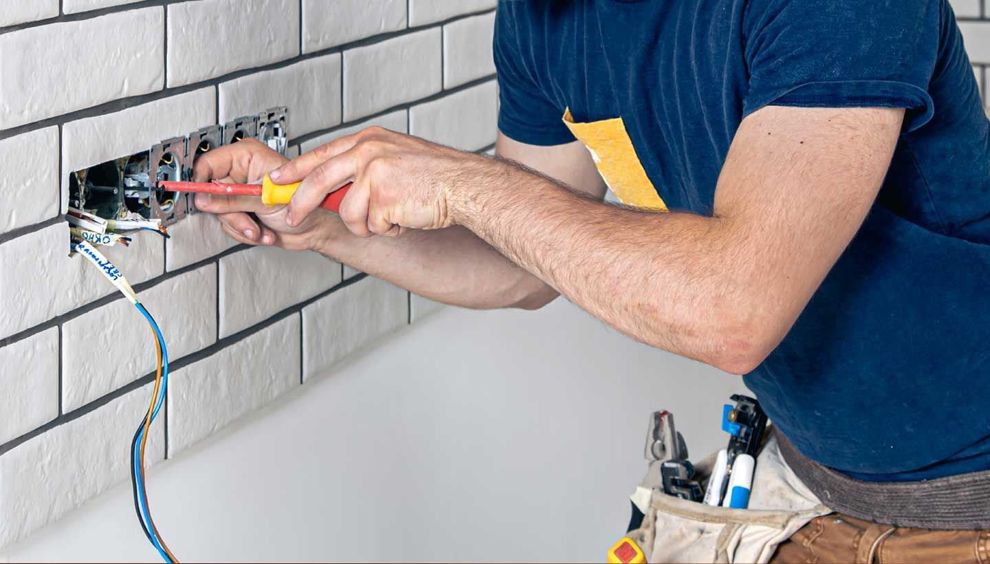 Electrician installing an electrical outlet on a white brick wall, using a screwdriver.