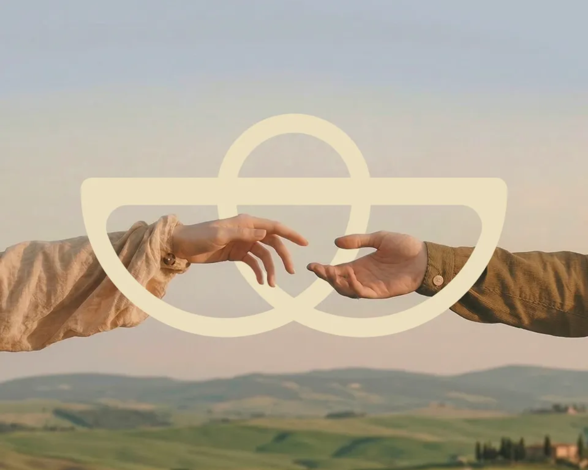 Two hands reaching toward each other over a soft, cream-colored symbol above rolling green hills