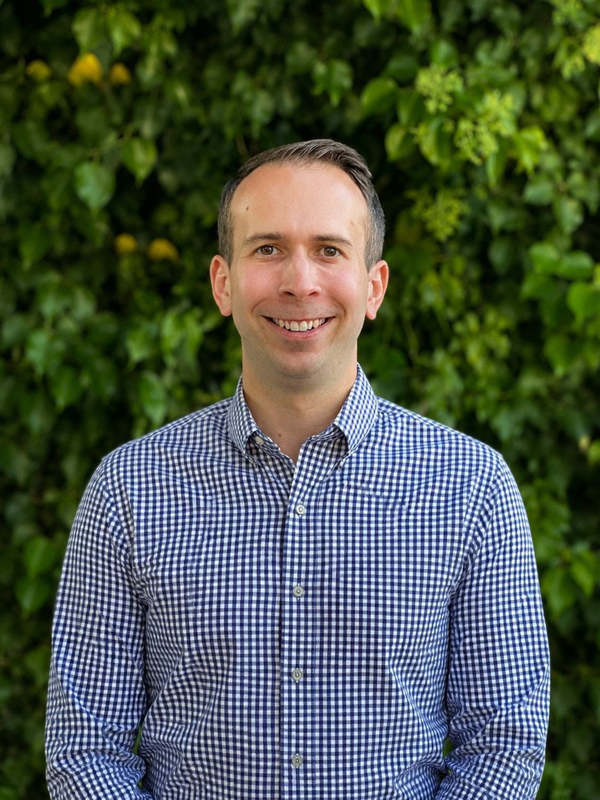 Dr. Charles William Dickey smiling in a blue checkered shirt standing outdoors against green foliage