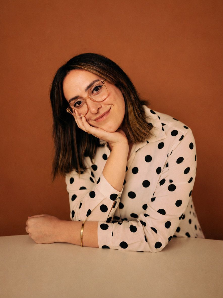 Dr. Elika Ramzjou in a white polka-dot shirt sitting on a couch by a sunny window, smiling indoors