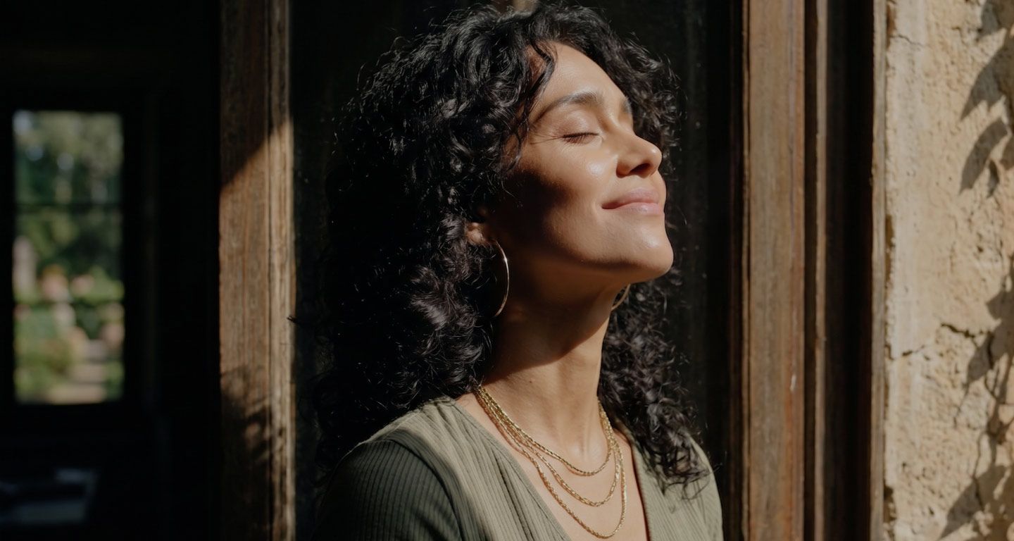 Woman with curly hair basking in sunlight by a stone wall, eyes closed and smiling softly.