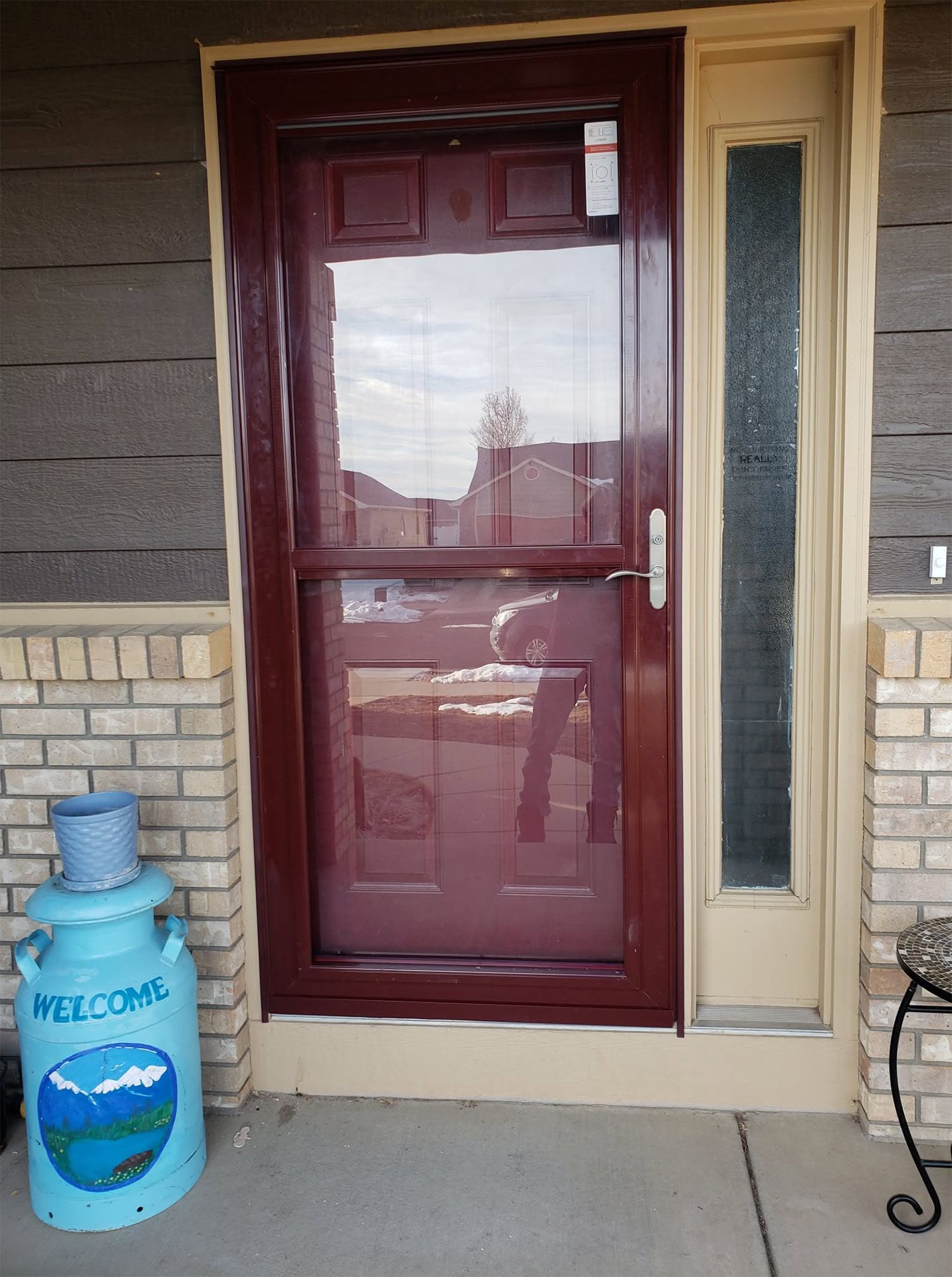 Front porch with burgundy storm door, narrow side window, and blue “Welcome” decorative bottle.