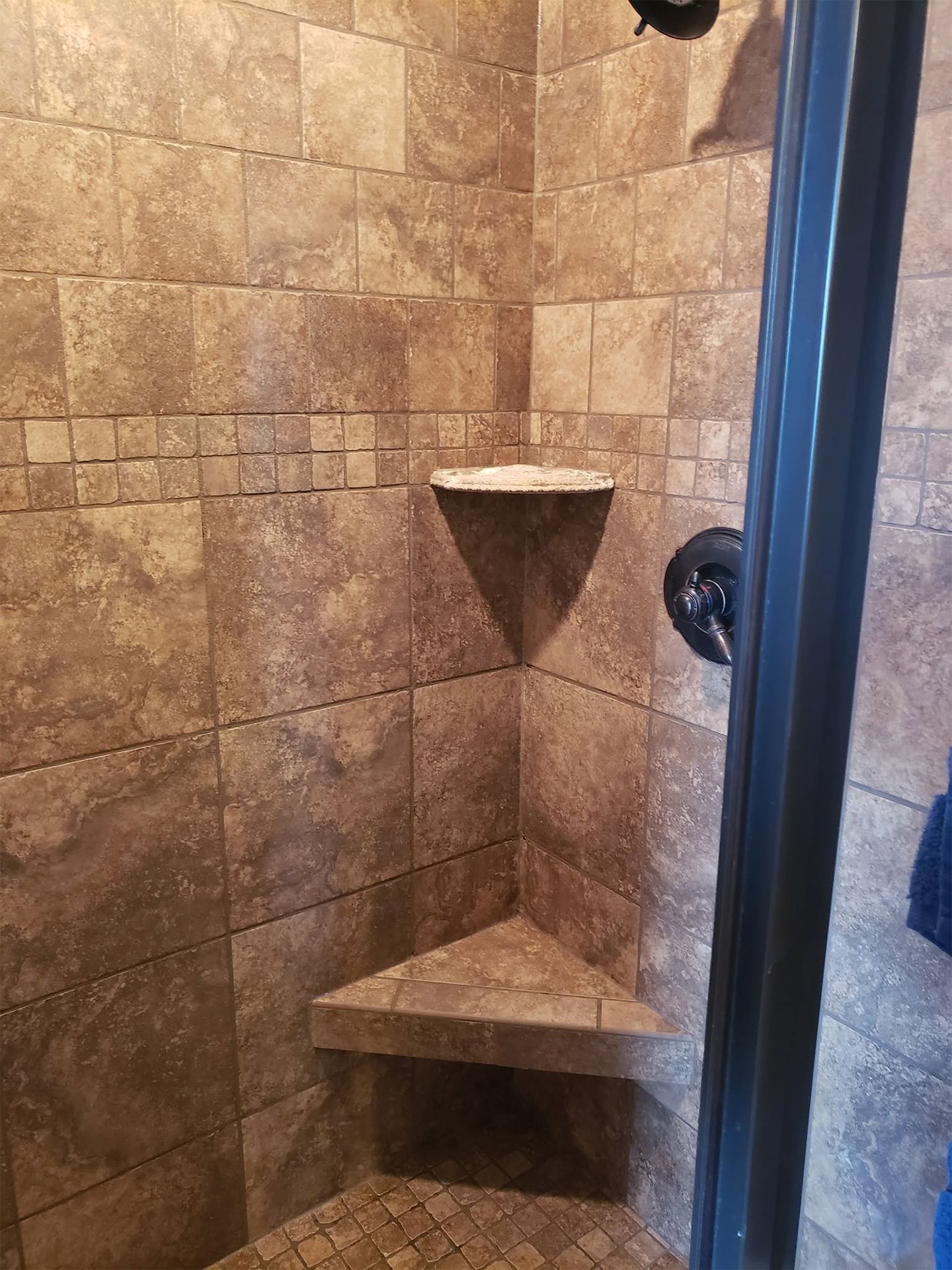 Stone-tiled shower stall with corner shelves and a glass door handle.