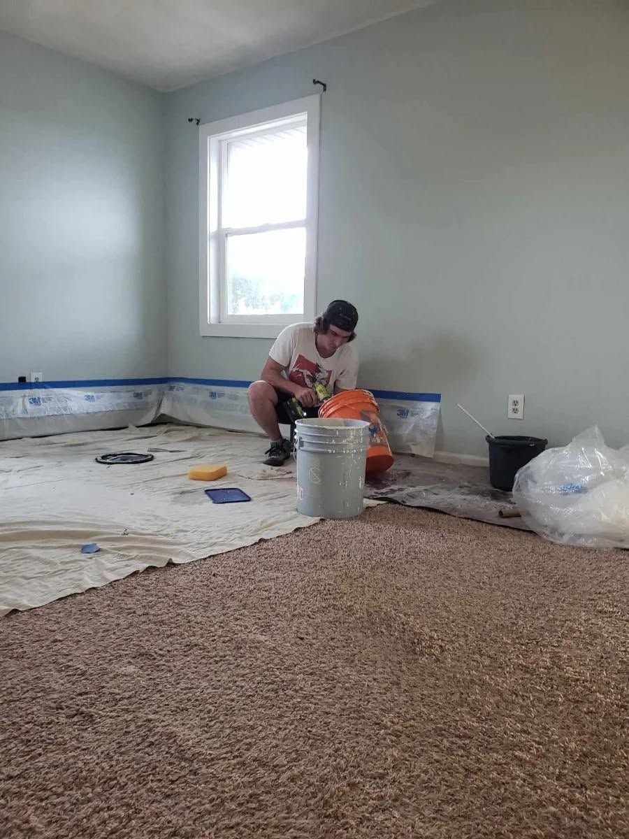 Person installing carpet in a mostly empty room with gray walls and a window.