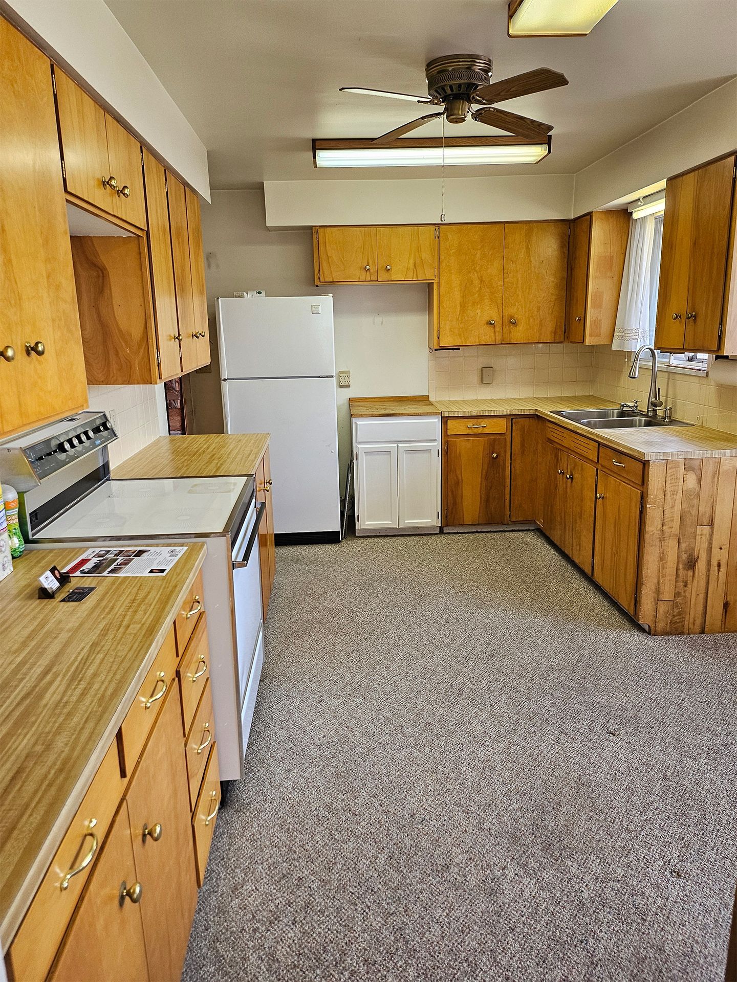 Small vintage kitchen with wood cabinets, white appliances, speckled carpet, and a ceiling fan