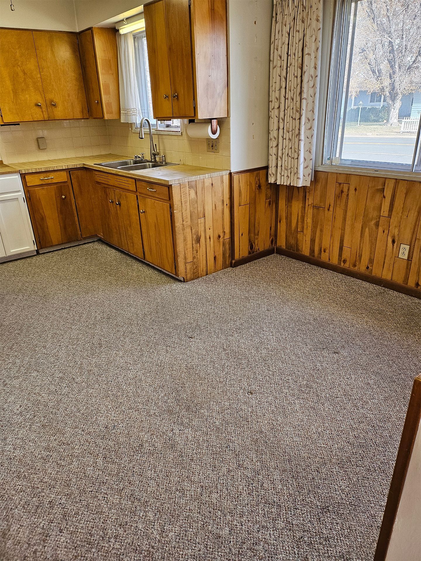 Empty kitchen with wood cabinets, beige carpet, and a window with curtains.