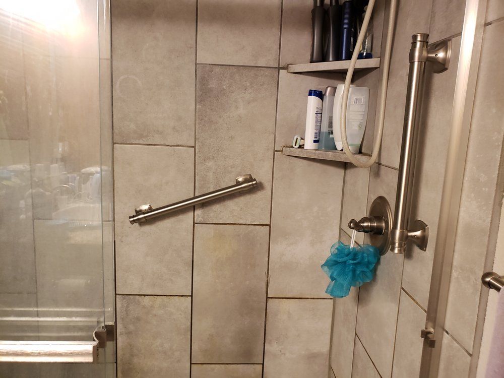 Walk-in shower with beige tile, glass door, metal grab bar, and blue loofah hanging on the faucet