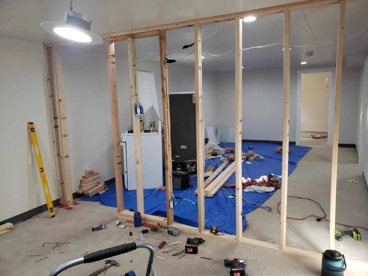Interior under renovation with exposed wall framing, blue tarp on floor, and construction tools scattered around.