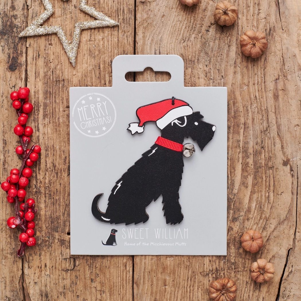 Kerstversiering voor een hond: een zwarte schnauzer met een kerstmuts en -halsband. Grijze kaart op hout met versieringen.