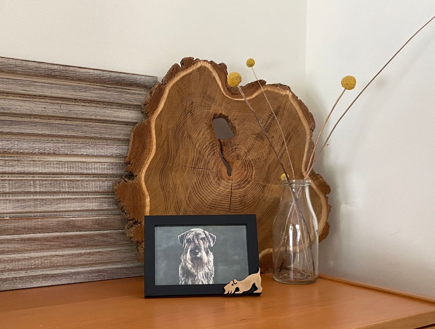 Hoekdisplay: houten plaat, ingelijste foto van een hond, glazen fles met gele bloemen.