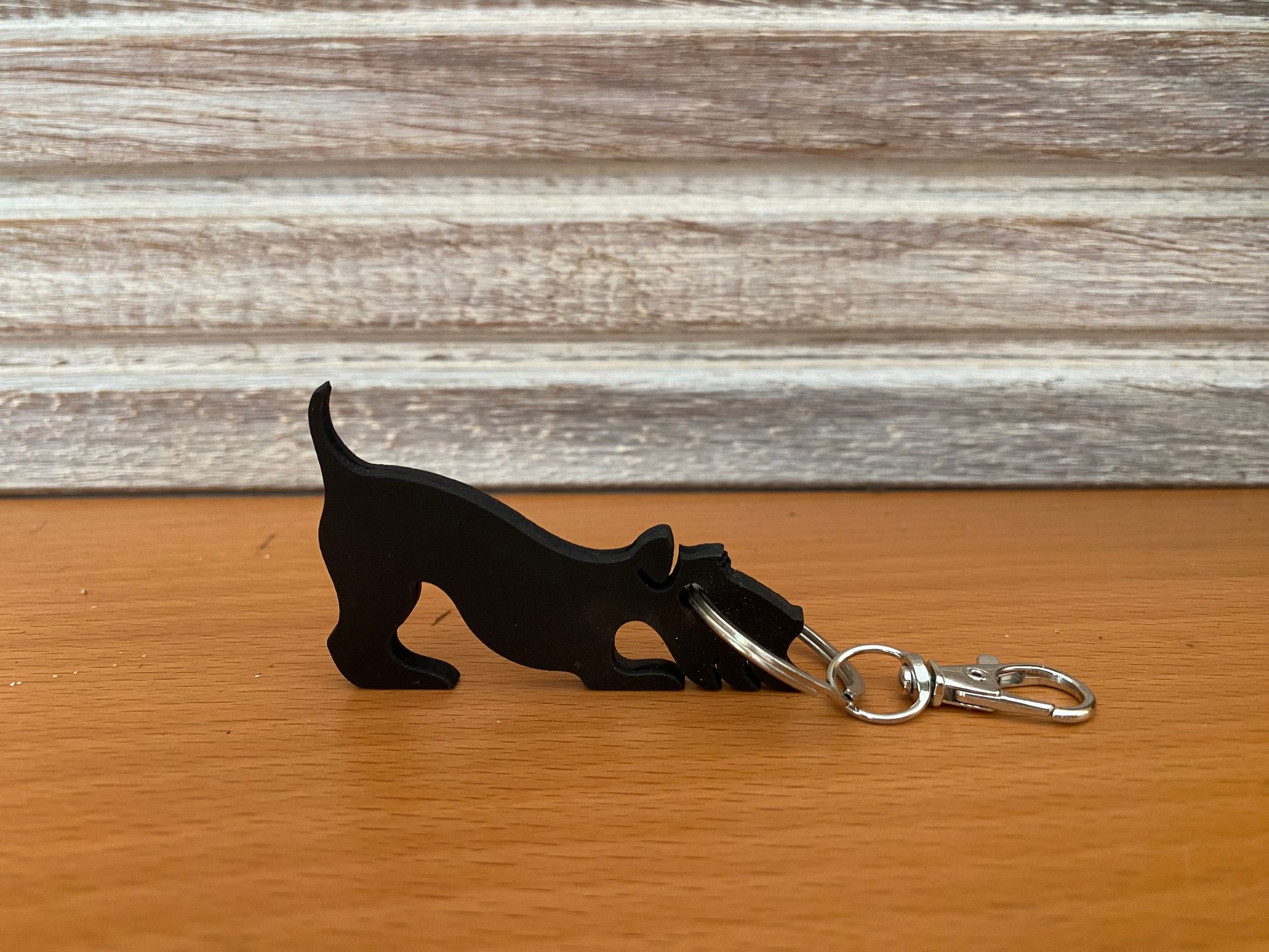 Sleutelhanger van een zwarte hond op een houten oppervlak, in een speelse houding met opgeheven staart.