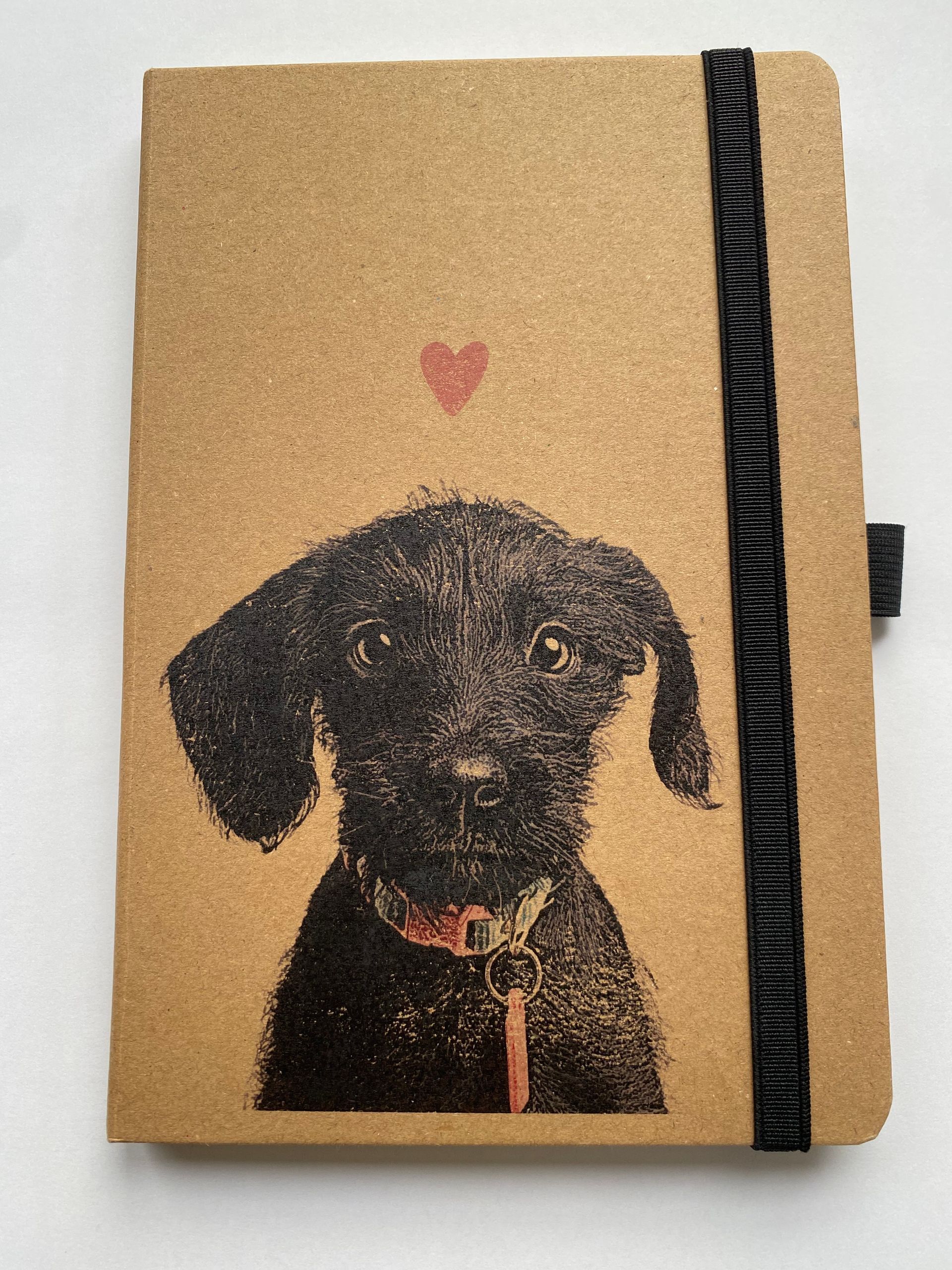 Notitieboekje met illustratie van een zwarte hond, roze hart en zwarte elastische band op een bruine kaft.