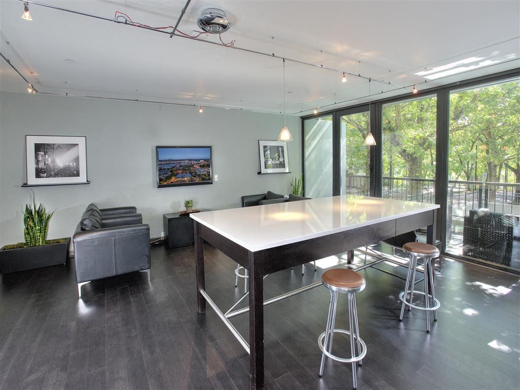 Modern community lounge with a high white table, stools, and large glass doors to a balcony at Harrison Tower