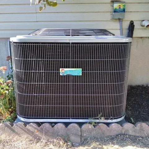 Outdoor air conditioning unit with black grille, mounted near a house.