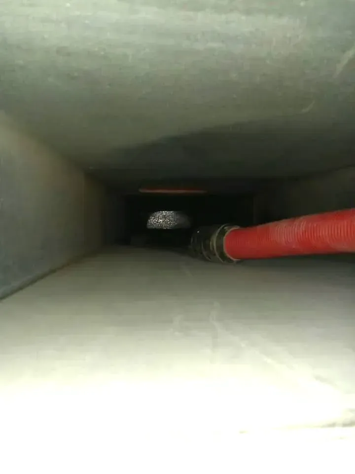 Inside of a dusty rectangular air duct, a red hose extends into the darkness.