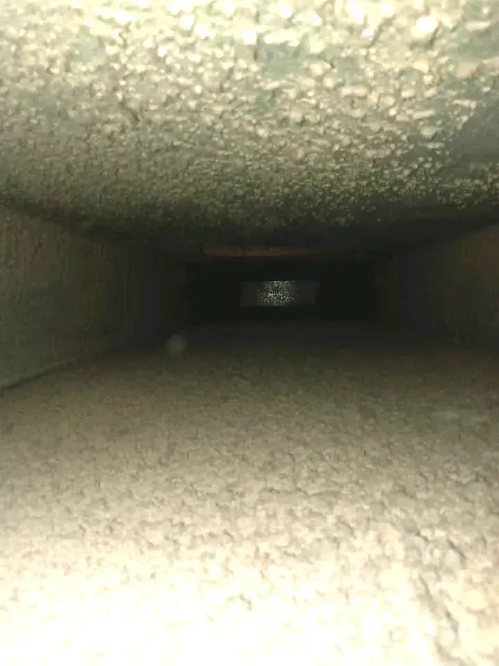 Dirty, rectangular HVAC duct interior. Dusty, light-colored debris coats the walls and floor, fading to darkness in the distance.