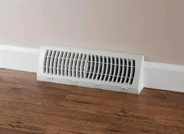 White rectangular floor vent against a beige wall and white baseboard, set in wood flooring.