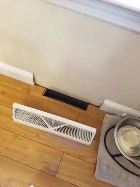 White vent cover on wood floor next to a black vent and a wall.