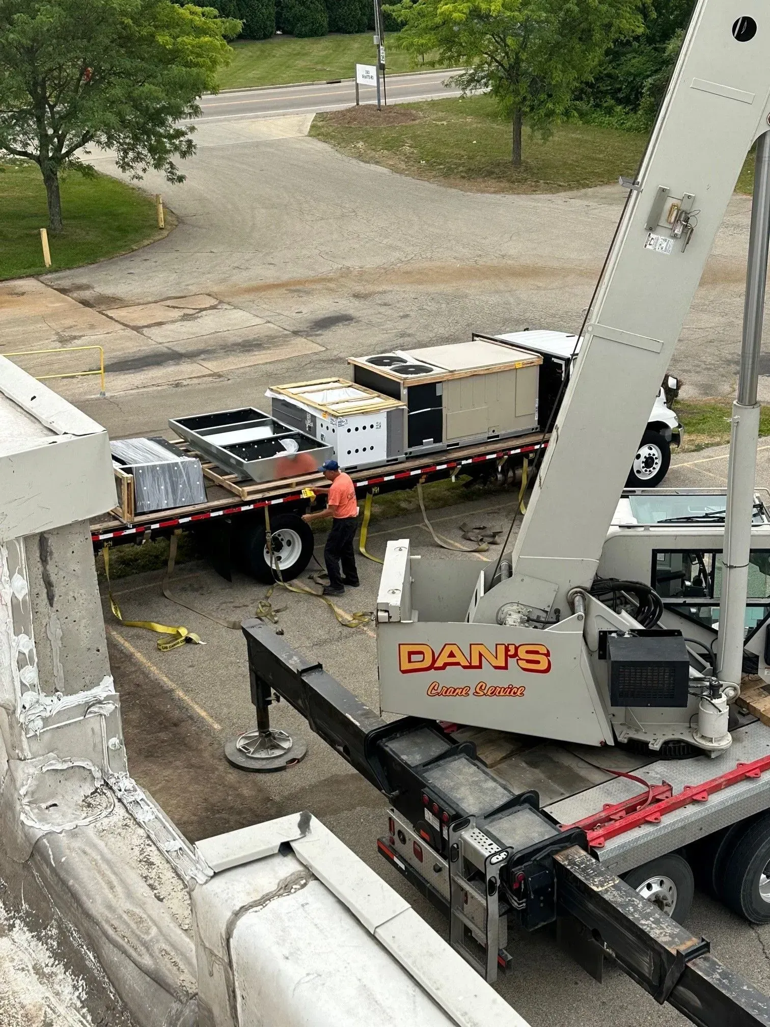 A crane lifts HVAC units from a flatbed truck. A worker in an orange shirt is present.