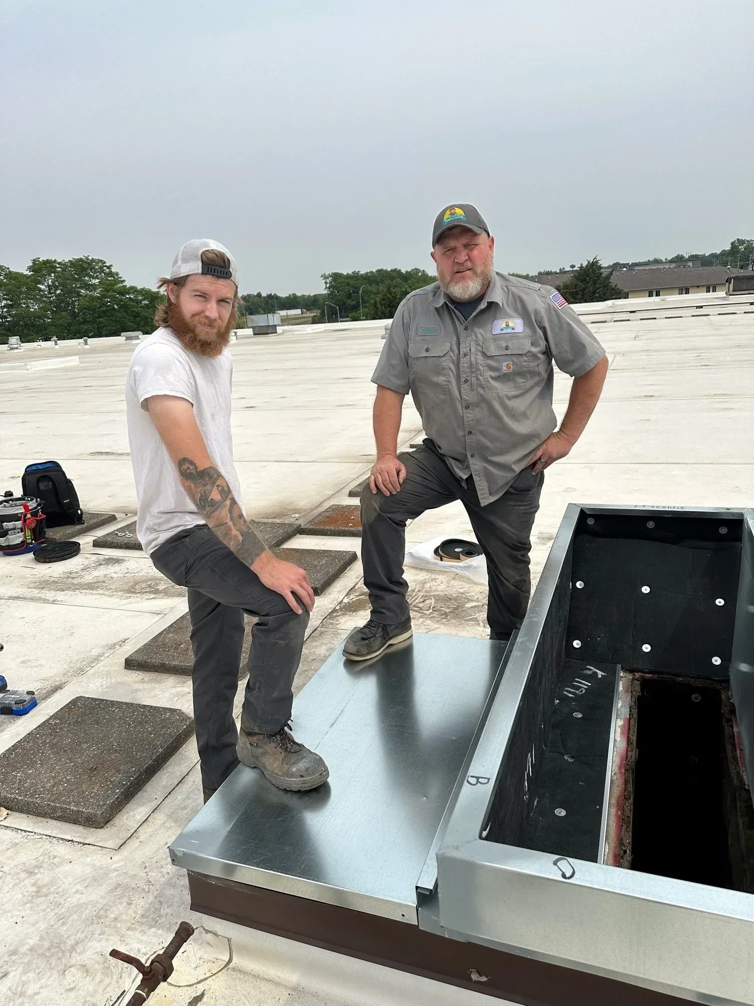 Two men on a flat roof next to a metal fixture, with tools scattered around. Cloudy sky.