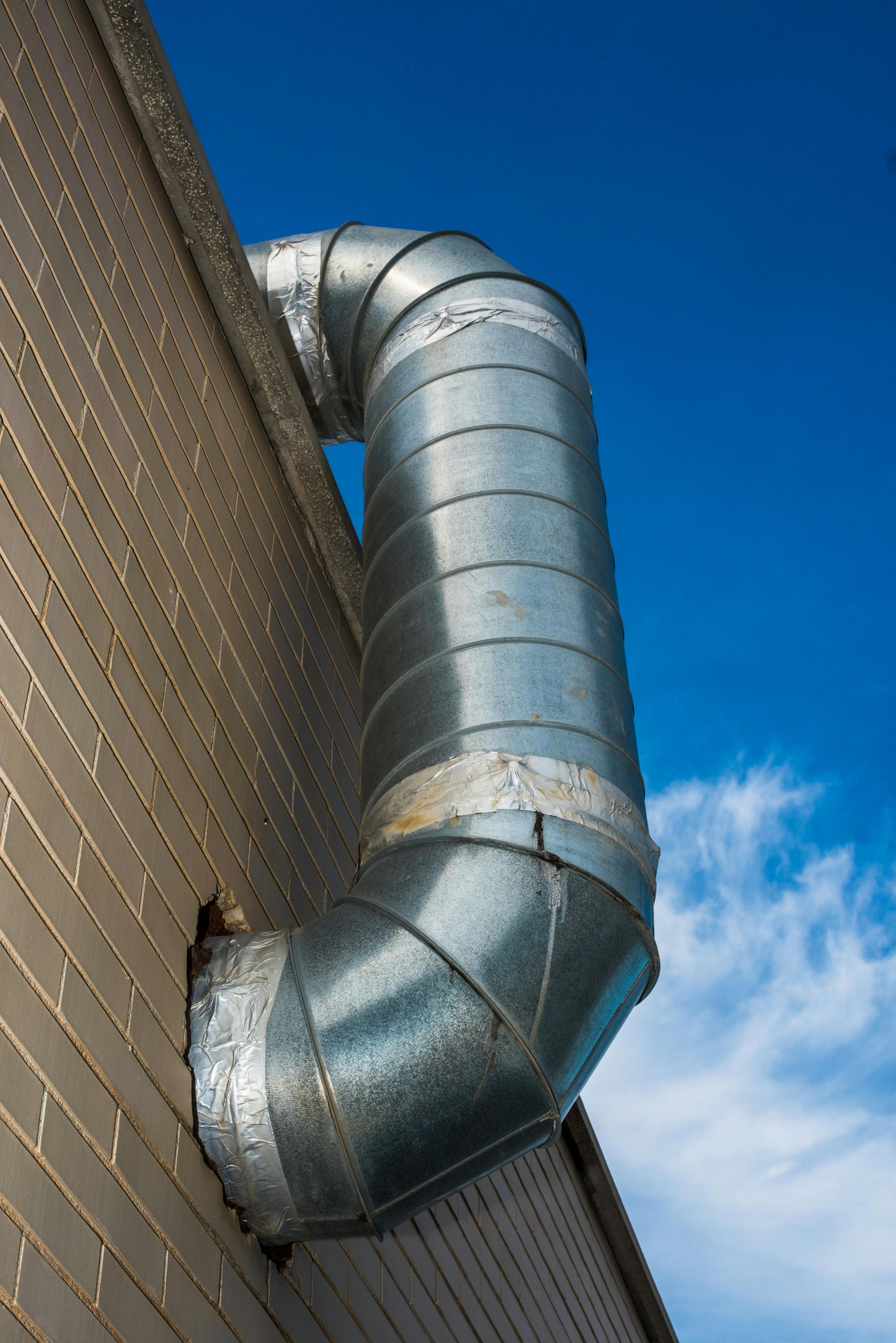 Metal ventilation ducting extends from a brick building, bending against a blue sky.