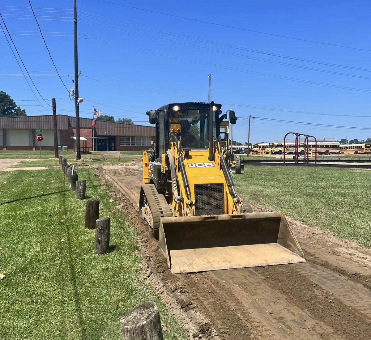 Yellow backhoe on a dirt path in a grassy area near a school, under a clear, blue sky.