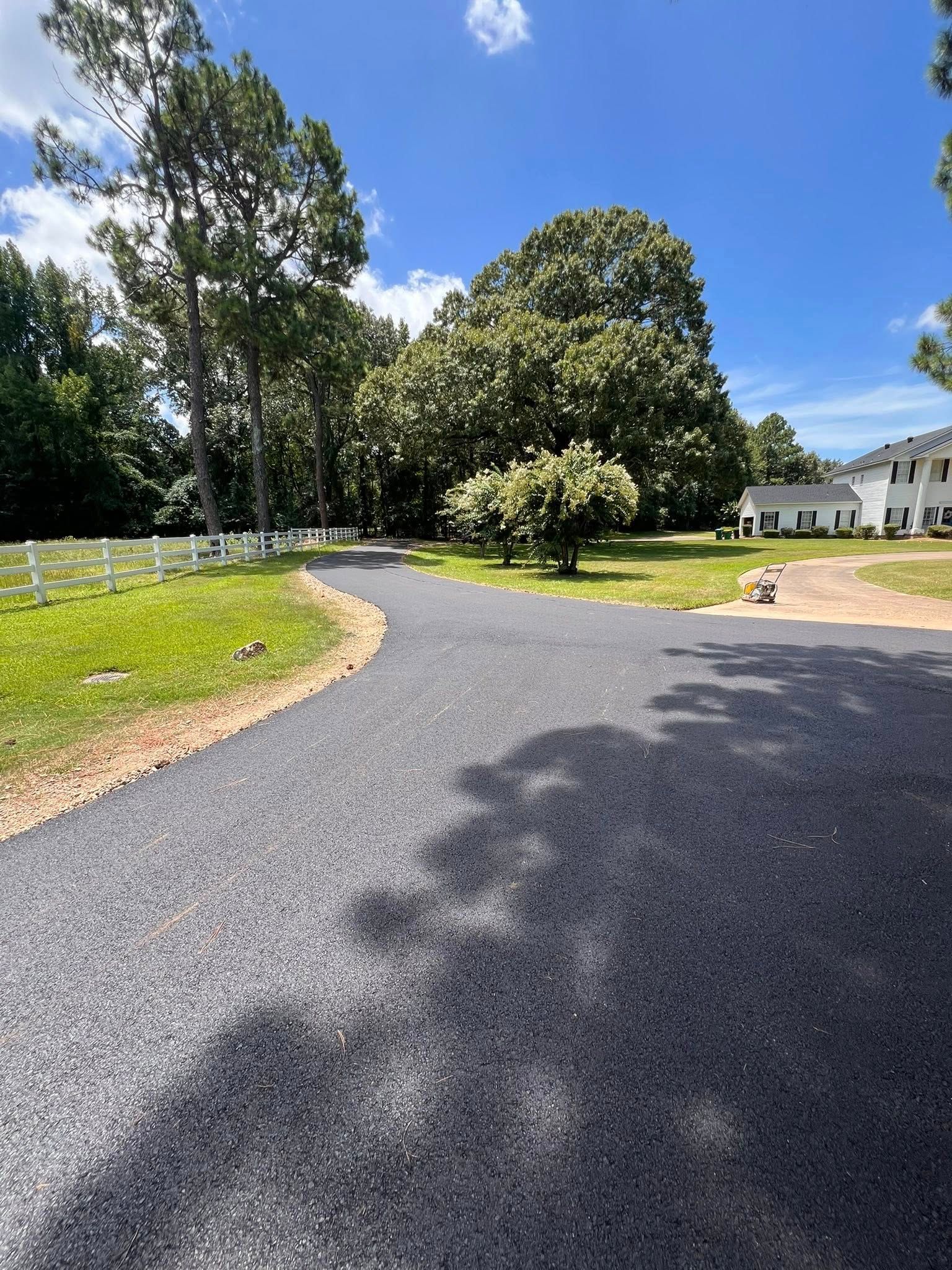 Newly paved dark gray driveway curves toward trees and white buildings under a blue sky.
