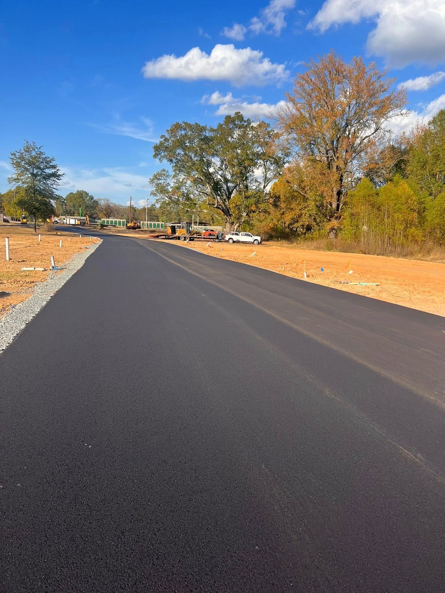 Newly paved asphalt road leads through a construction site, with trees and a blue sky overhead.