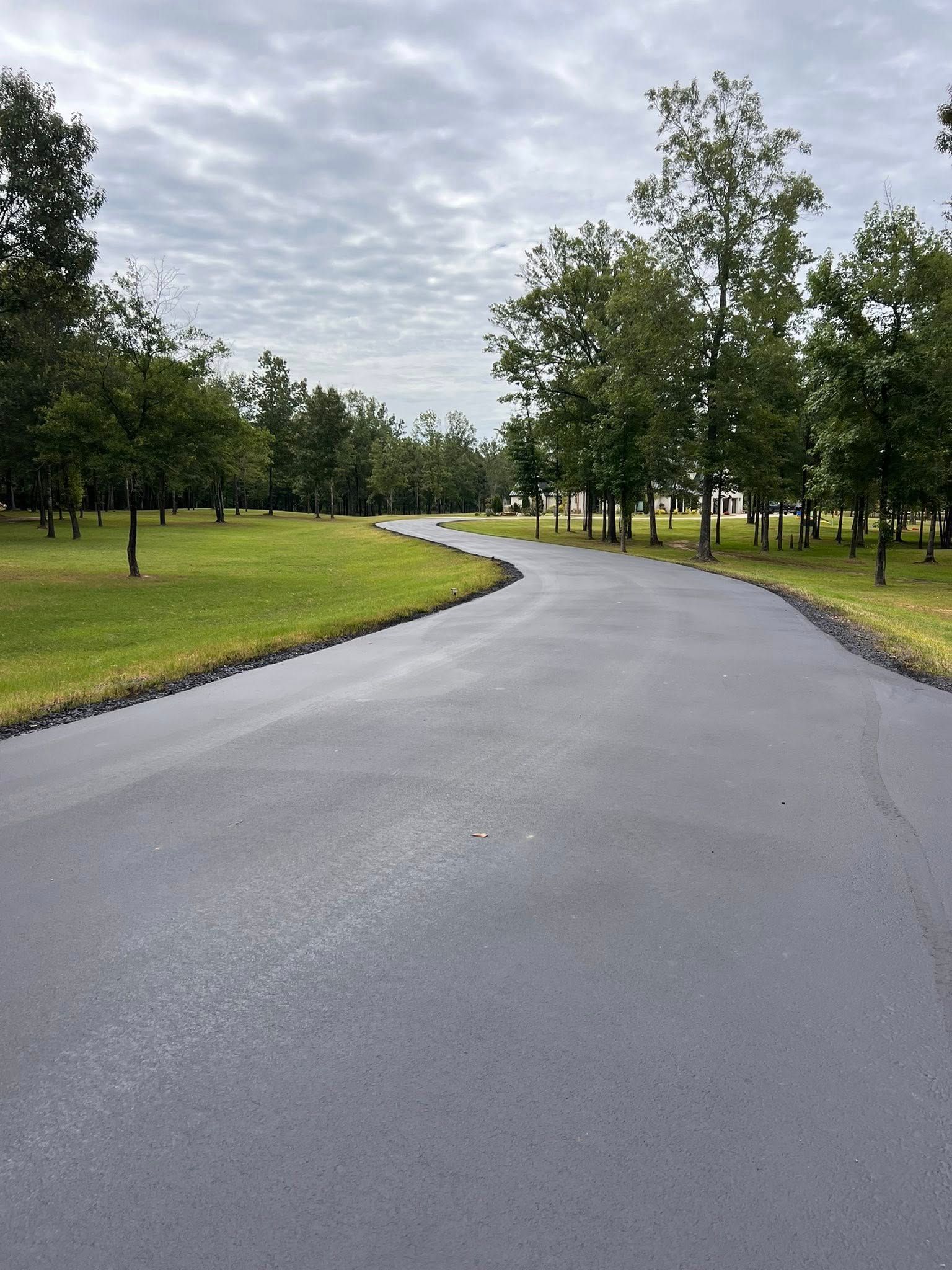 Black asphalt road winds through a grassy park lined with trees under a cloudy sky.