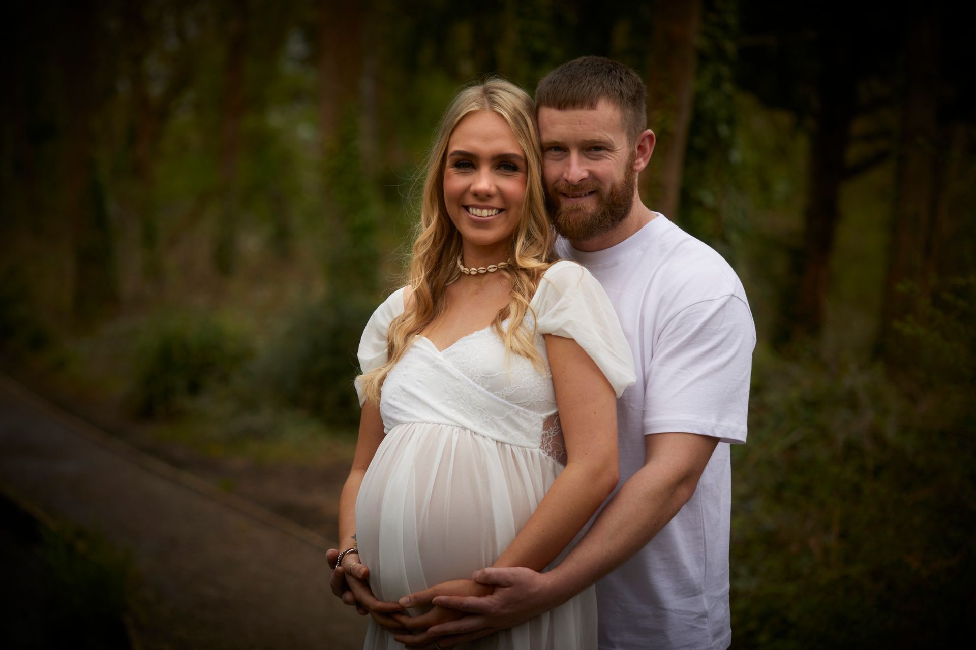 Pregnant couple embracing outdoors in a wooded setting, smiling at the camera
