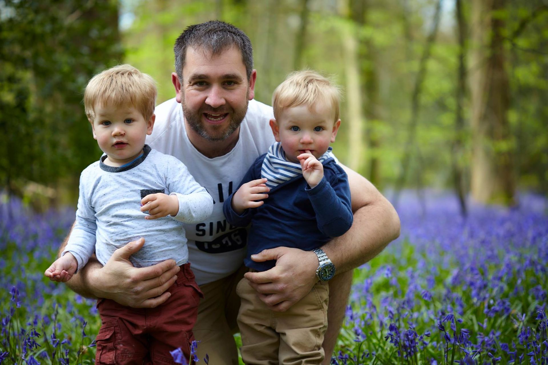 Man kneels in a purple flower field, holding two small children outdoors among trees.