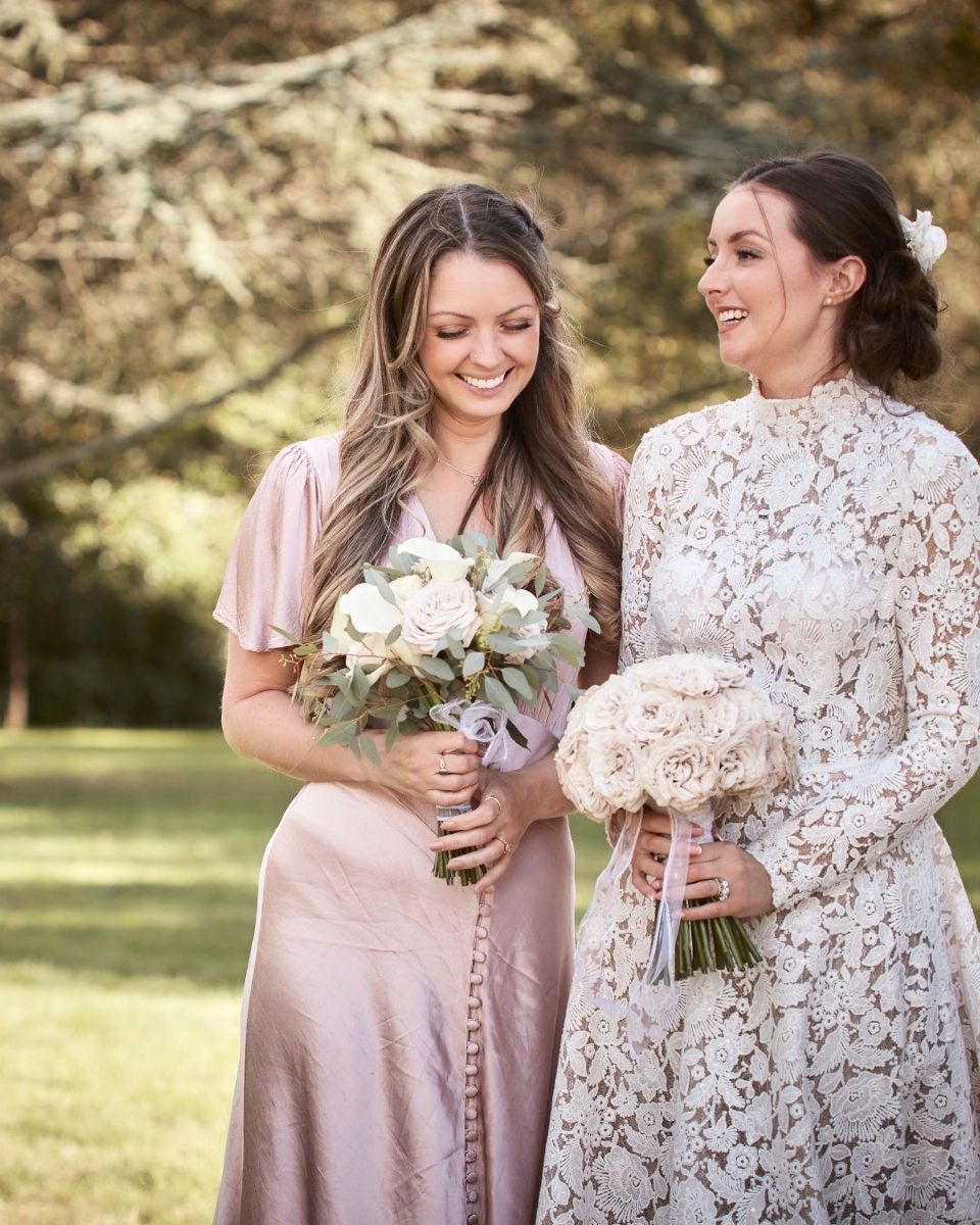 Two women in pastel dresses smiling outdoors, each holding a bouquet at a garden wedding