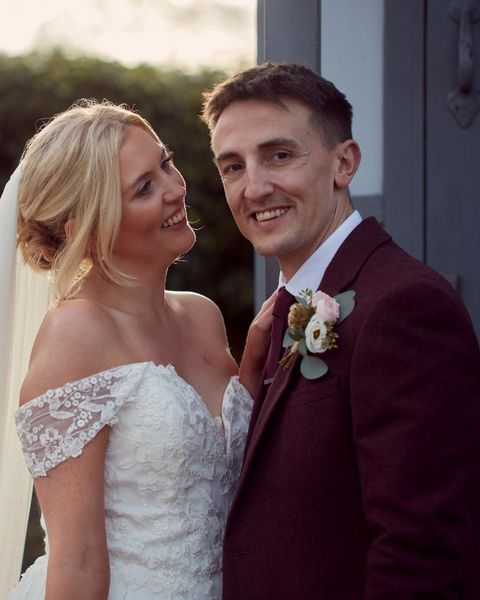 Newlywed couple smiling outdoors, bride in white lace dress and groom in burgundy suit