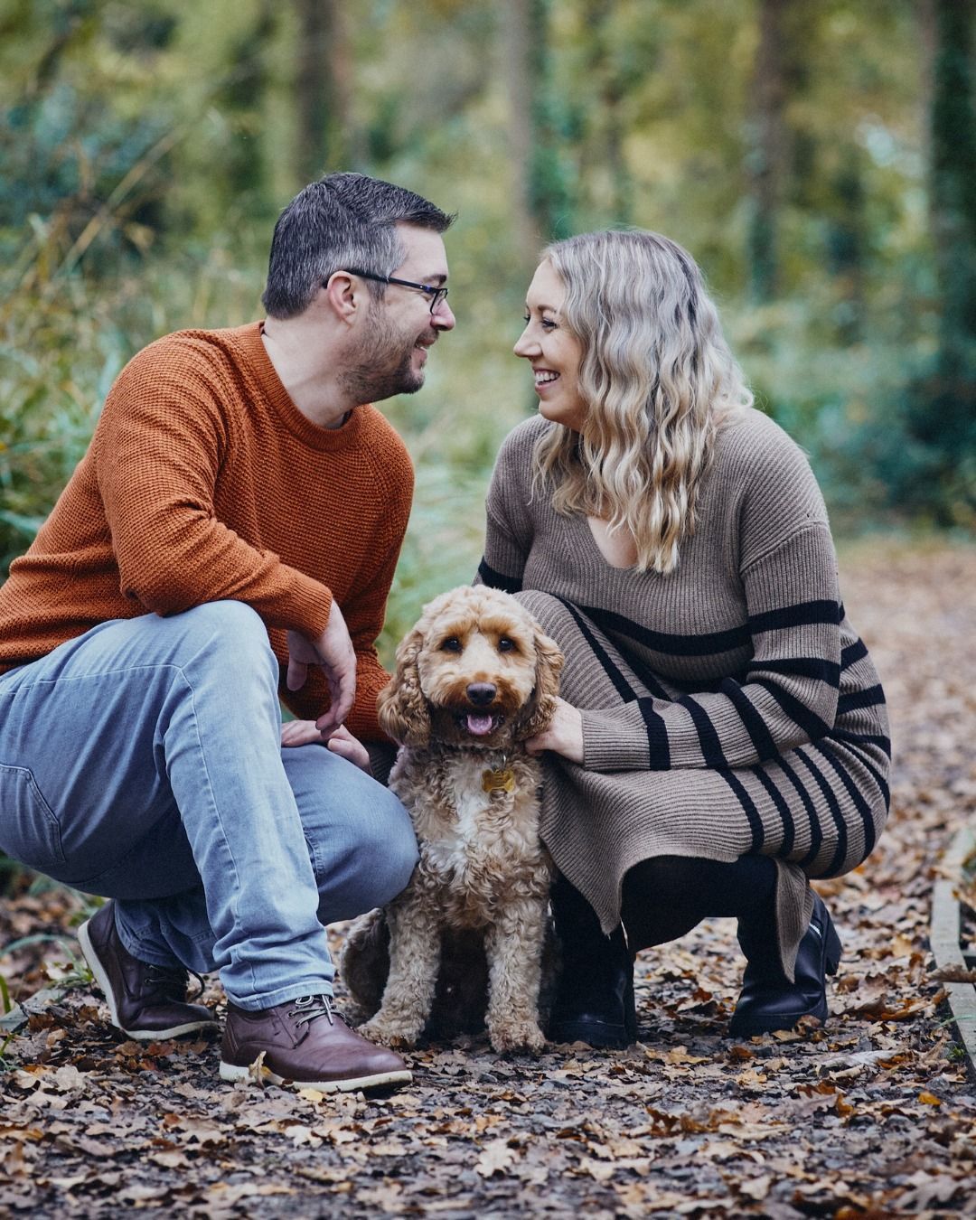 Couple crouches with a curly-haired dog in a wooded outdoor setting, smiling at each other.