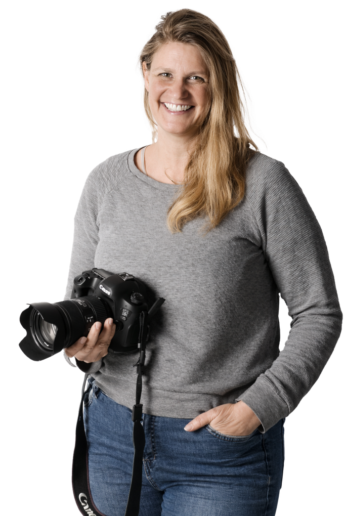 Woman holding a camera, wearing a gray sweater and jeans, smiling against a white background