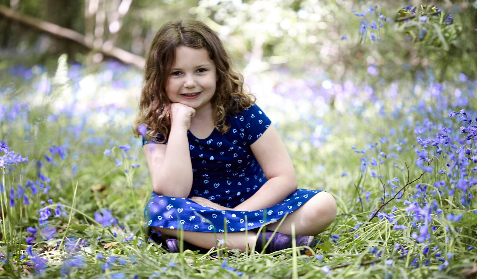 Young child in a blue polka-dot dress sitting in grass with purple flowers, smiling at the camera