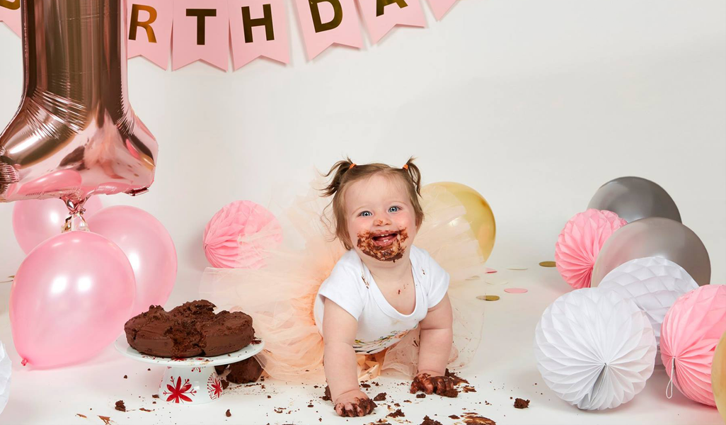 Toddler crawling through a pink birthday party setup with balloons, cake, and confetti.