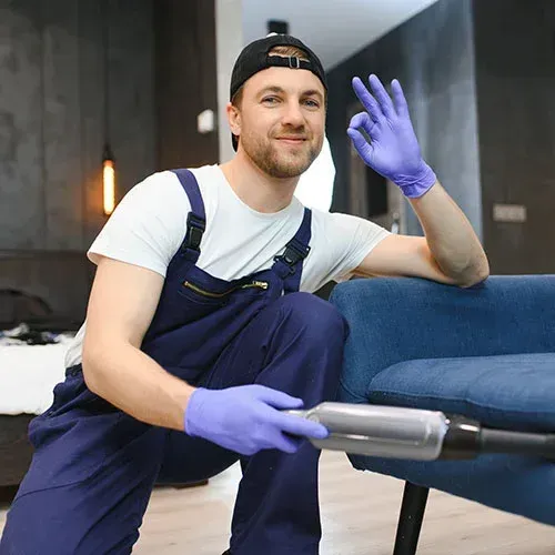 Man in overalls and gloves cleaning a blue couch, giving an