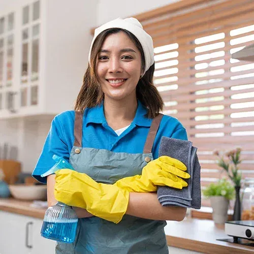 Woman in cleaning uniform smiling, holding spray bottle and cloths in a kitchen.