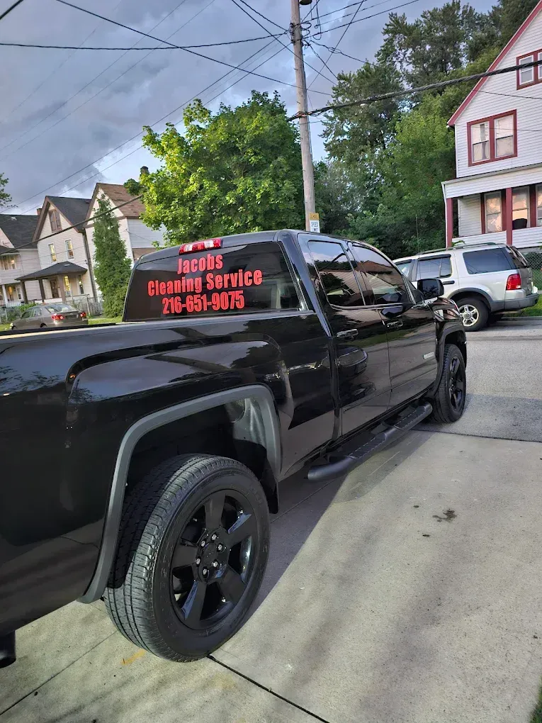 Black pickup truck parked on a driveway with business advertisement on back window. Houses and trees are in the background.