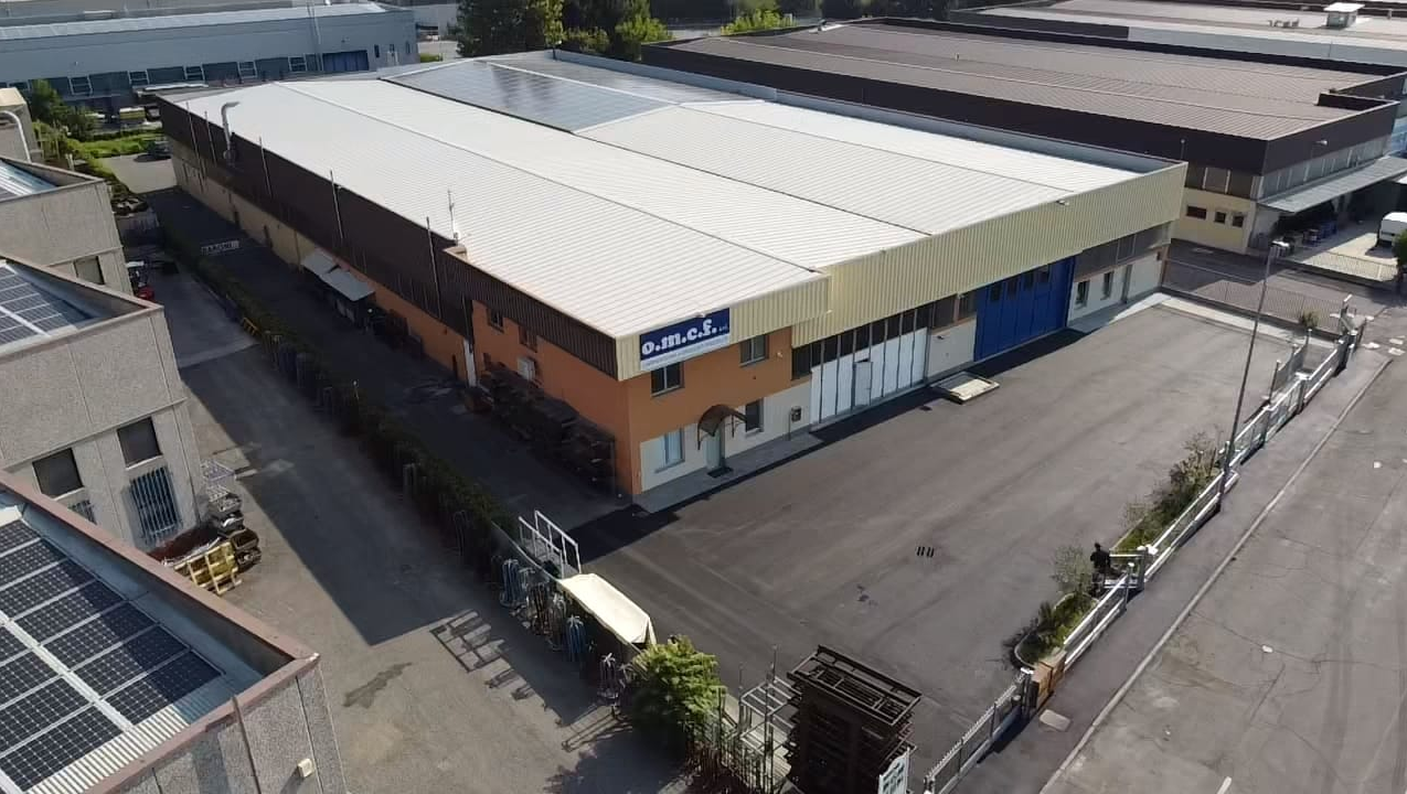 Veduta aerea di un grande magazzino industriale con tetto bianco e piazzale pavimentato vuoto.