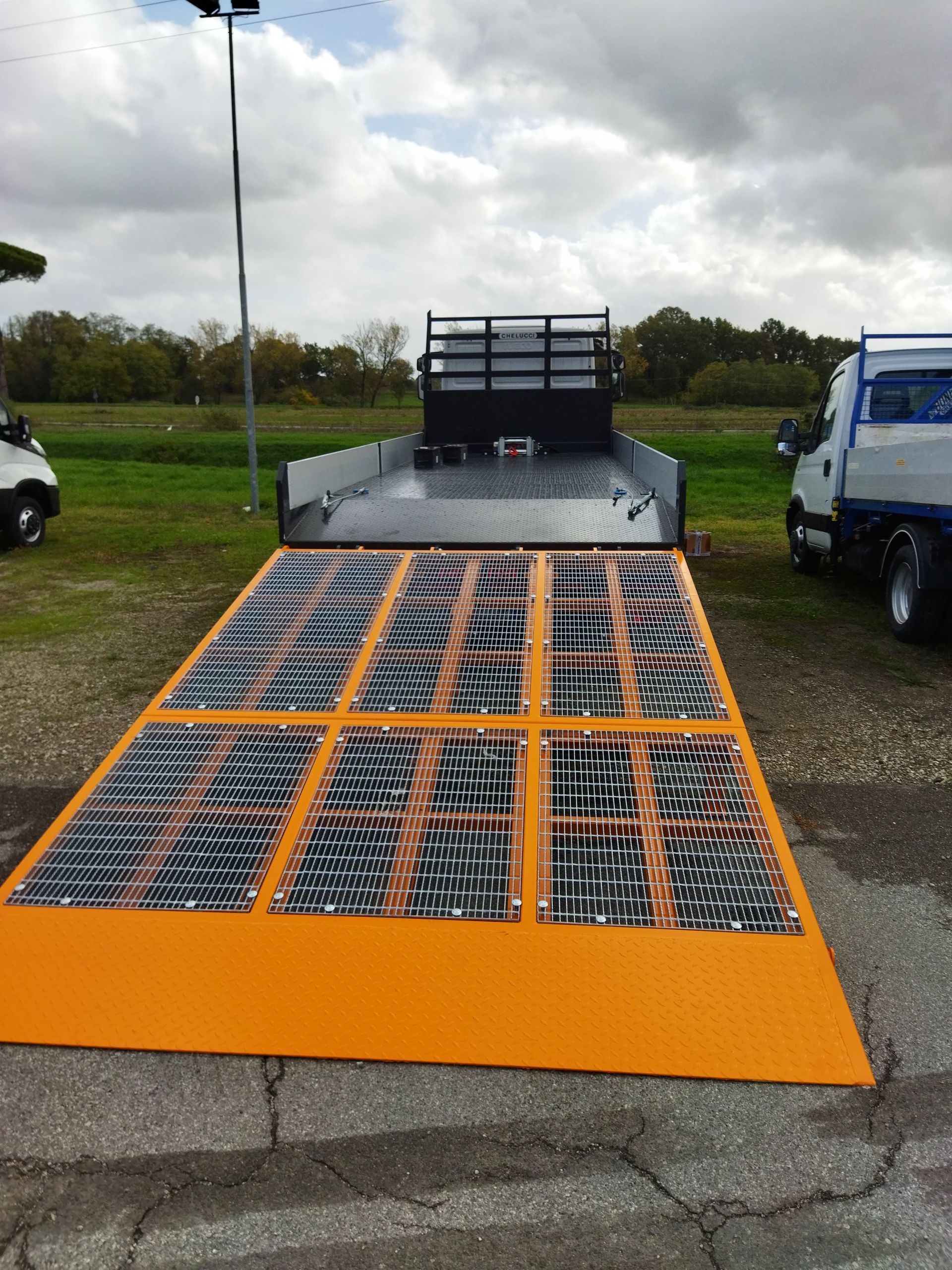 Rampa arancione con griglie metalliche che conduce al cassone di un camion in un parcheggio esterno nuvoloso.