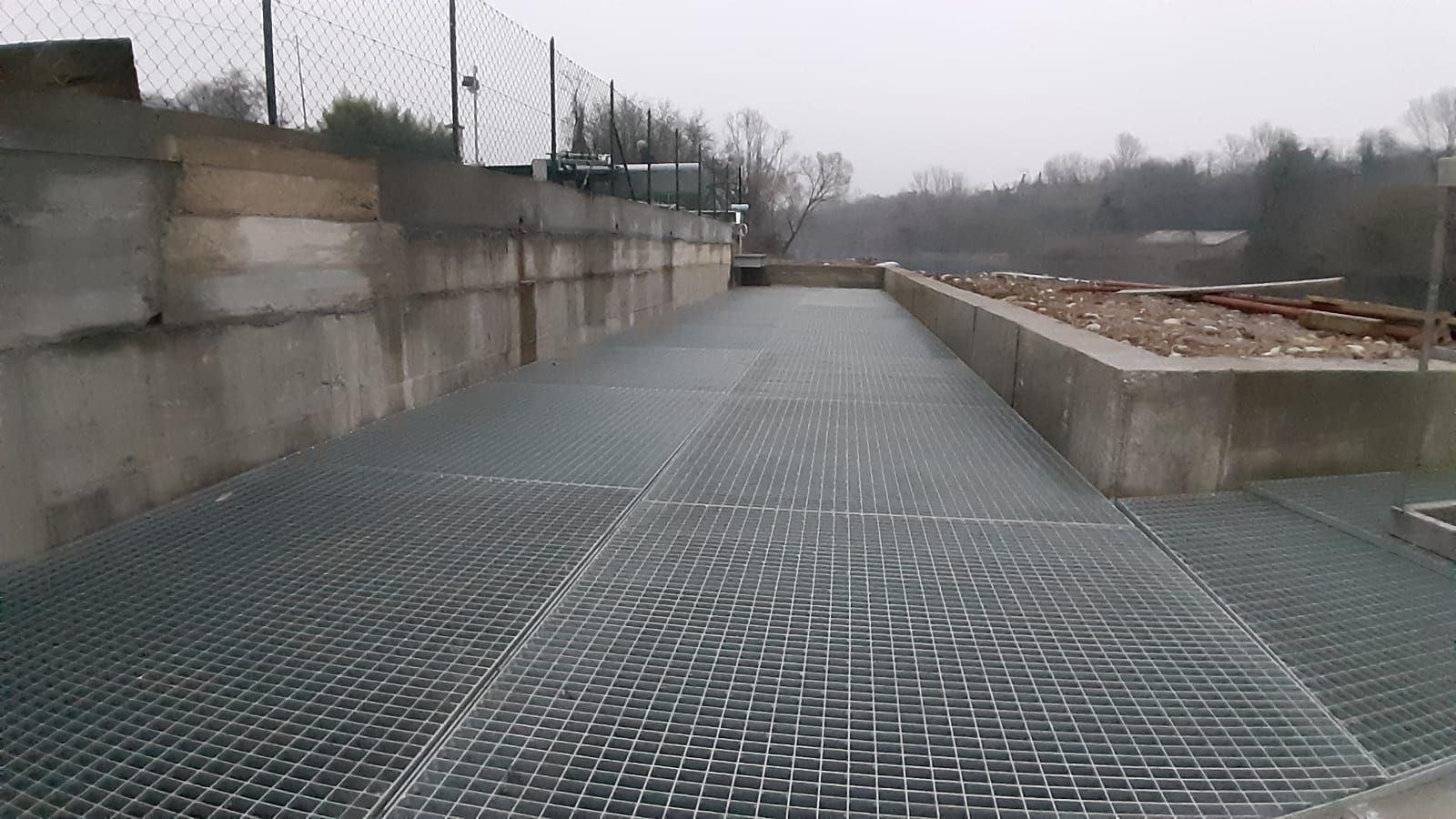 Una passerella in grigliato metallico accanto a muri di cemento e una recinzione in una giornata grigia e nuvolosa.