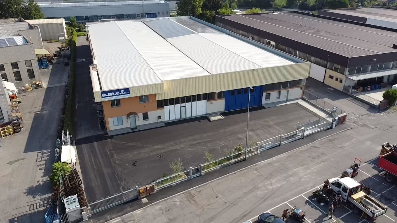 Veduta aerea di un grande magazzino con tetto bianco, situato accanto a una strada industriale e a un parcheggio.