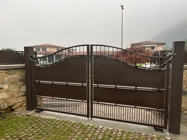 Cancello carrabile in metallo marrone con parte superiore ad arco decorativo davanti a un muro di pietra e case