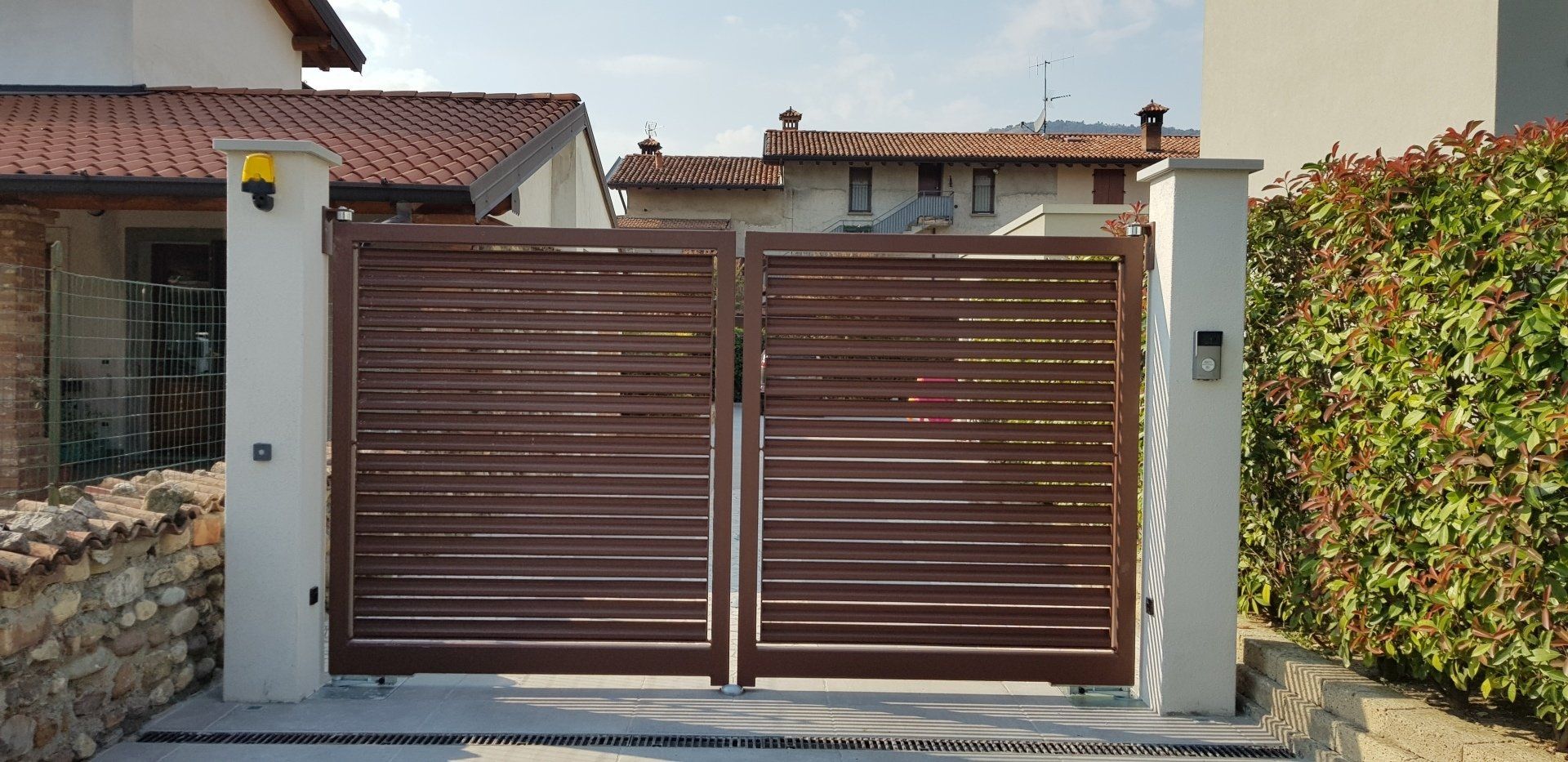 Doppio cancello metallico marrone davanti a una casa, fiancheggiato da pilastri in cemento e siepi.