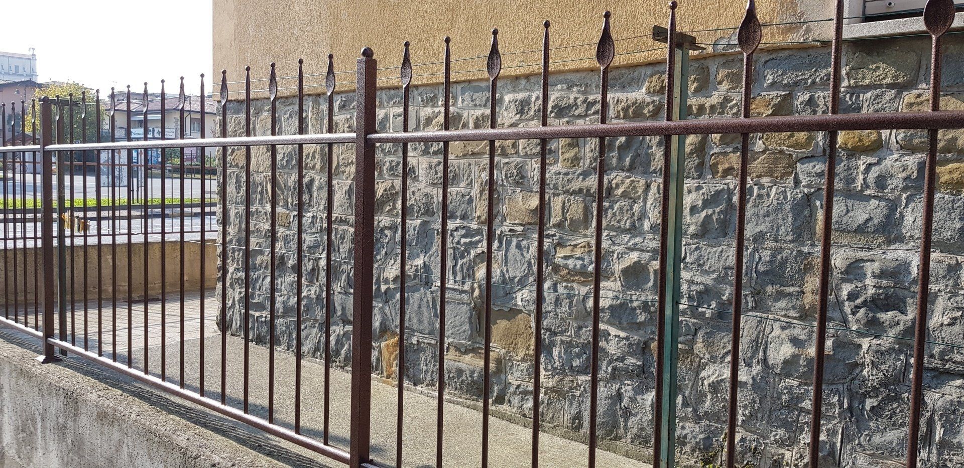 Recinzione metallica nera davanti a un muro di contenimento in pietra, accanto a un vialetto pavimentato.