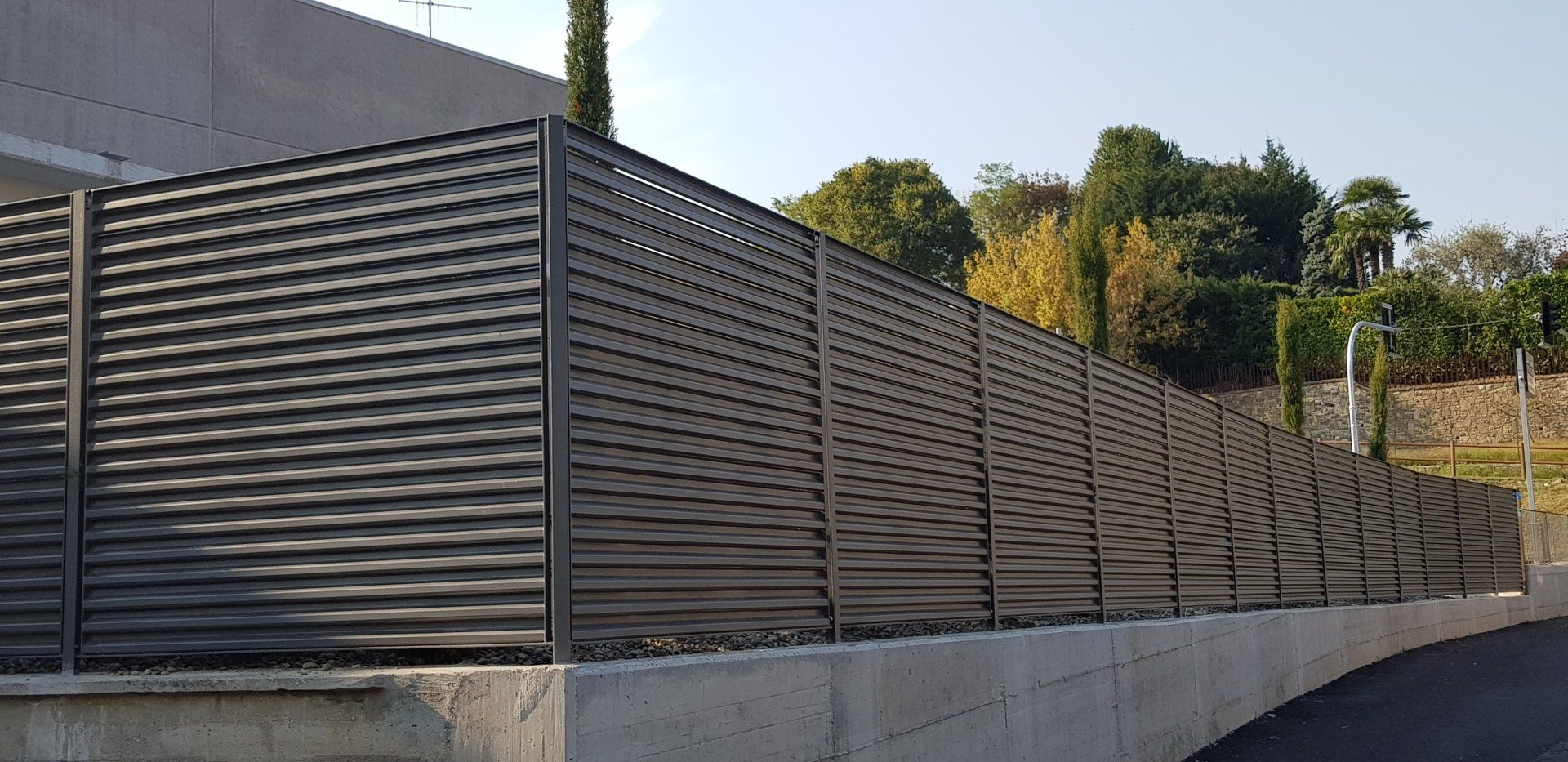 Recinzione a doghe grigie lungo un muro di contenimento curvo in cemento a lato di una strada, con alberi sullo sfondo.