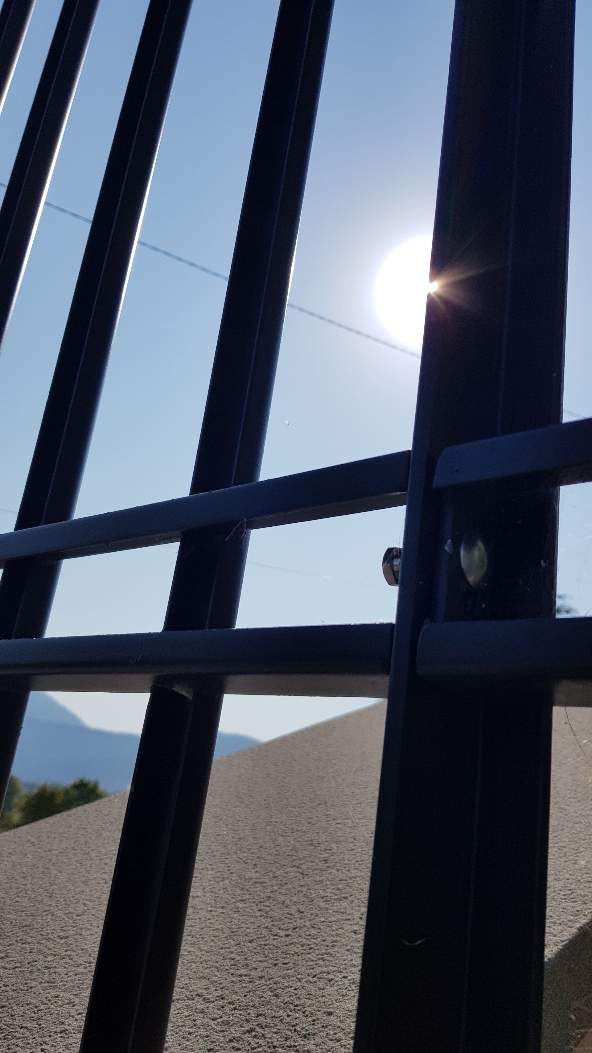 Luce solare filtra attraverso scure sbarre di metallo con sullo sfondo una collina desertica avvolta nella foschia.